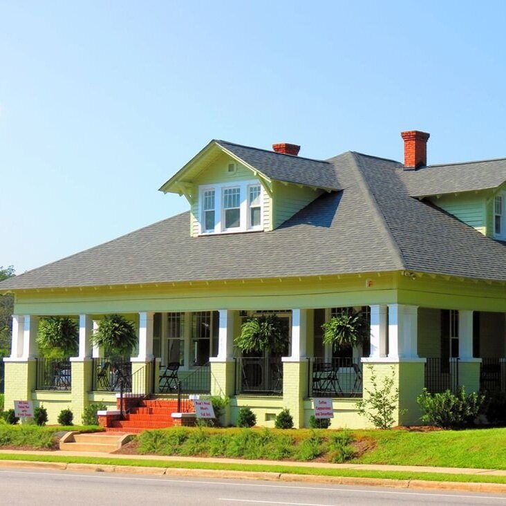 1495952_574623465947923_551675499_o Charming green Victorian-style house with a spacious front porch, featuring potted plants and inviting seating, located on a sunny street.